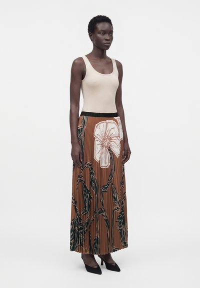 Femme portant un haut beige sans manches avec une jupe plissée marron taille haute ornée de grands motifs floraux blancs et verts et des talons noirs.