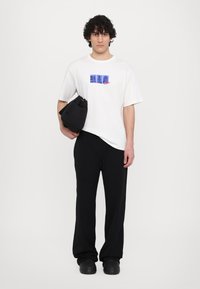 Λευκό t-shirt με μπλέ γραφικό σχέδιο, μαύρο παντελόνι σε φαρδιά γραμμή και μαύρη τσάντα. Απαλή ύφανση με χαλαρή εφαρμογή. Απλή φόντο.