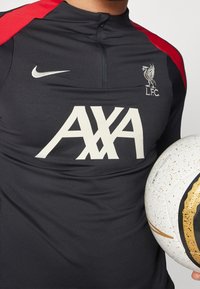 Schwarzes Sportshirt mit roten Akzenten, das über einen hohen Kragen, das Nike-Logo und großen weißen AXA-Text verfügt. Ein gemusterter Fußball wird in der Nähe gehalten.