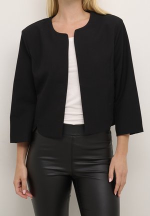 Femme portant une veste noire sans col, ouverte sur le devant avec des manches trois-quarts, par-dessus un haut blanc et un pantalon en cuir noir.