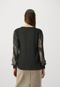 Svart bluse med gjennomsiktige blonde ermer med prikkete mønster og volanger. Enkel rund halslinje og tettsittende kroppsdesign.