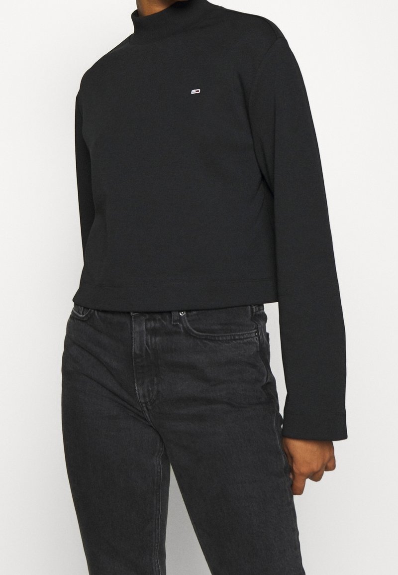 Svart kort långärmad topp med hög hals, tillverkad av ett slätt tyg, med en liten logotyp på bröstet. Kombinerad med svarta jeans.