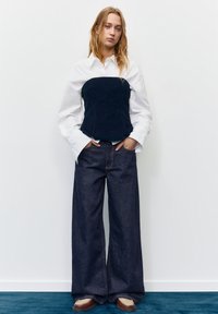 Mlada ženska z razpuščenimi blond lasmi, oblečena v belo majico z dolgimi rokavi, črno majico brez rokavov, široke temne kavbojke in rjave čevlje, stoji na modrem preprogi.