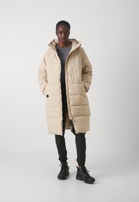 Manteau long beige en doudoune avec un motif matelassé horizontal, fermeture éclair et capuche. Associé à un pantalon noir et des chaussures de sport noires.