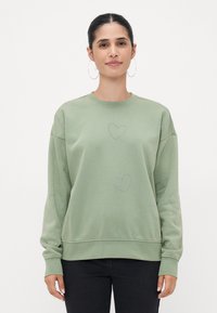 Svetlozeleni pulover iz mehkega blaga, z akcenti v obliki srčkov na sprednjem delu in rokavih, sproščenega kroja ter rebrastimi manšetami.