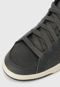 Sapatilha cinza escuro com parte superior em couro texturizado, fecho de atacadores, caixa dos dedos perfurada e sola de borracha branca. Os detalhes incluem costura visível.