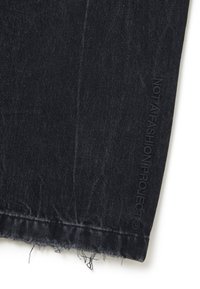 Tejido de mezclilla oscura, con dobladillo deshilachado y el texto en relieve "NO ES UN PROYECTO DE MODA" en el lado. Textura suave, corte de jeans estándar.