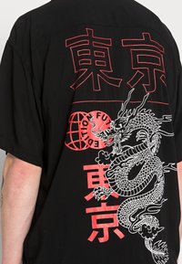 Camicia nera oversize con grafica di drago rosso e testo, con una texture morbida e maniche corte, che mette in evidenza dettagliate decorazioni.