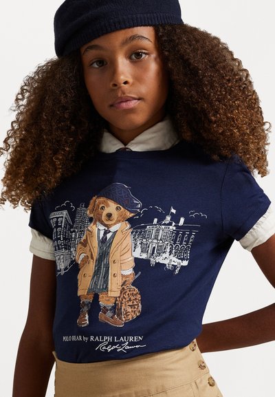 Camiseta de color navy que presenta un gráfico de un oso con abrigo y sombrero. La camiseta tiene mangas cortas y un escote redondo. Detalle del cuello en un color claro.
