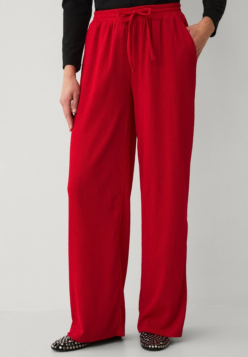 Vrouw draagt losse, rode broek met elastische tailleband en trekkoord, gecombineerd met zwarte lange mouwen en zwarte schoenen met zilveren studs.