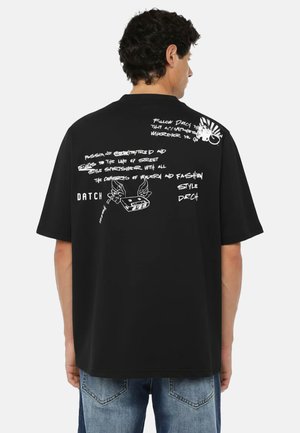 Uomo con capelli ricci che indossa una T-shirt nera oversize con testo bianco in stile graffiti e un disegno di una cassetta alata sul retro.