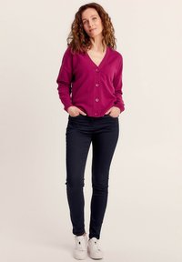 Cardigan fucsia con bottoni e scollo a V, polsini a coste e vestibilità rilassata, abbinato a jeans skinny in denim scuro e sneakers bianche.