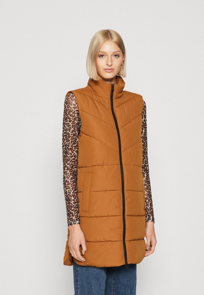 Brun padded vest med høy krage, front glidelås og skrå sømmer. Båret over en mønstrete skjorte med lange ermer og sammen med blå jeans.