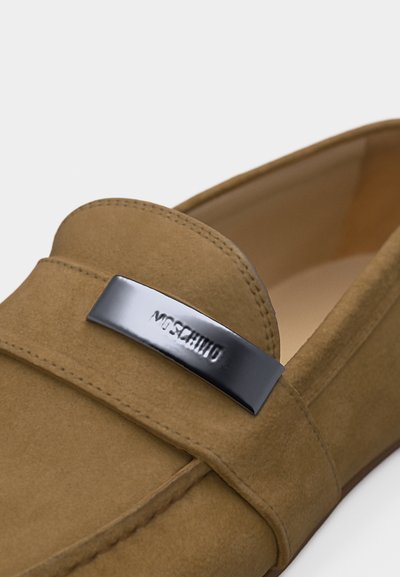 Mocassin en daim beige avec une plaque en métal argenté gravée de "MOSCHINO" sur la bride traversant le dessus du pied.
