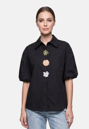 Femme portant une chemise noire à boutons avec des manches bouffantes et trois épingles décoratives en forme de fleur en vert, pêche et blanc sur le devant.