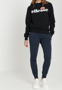 Sudadera negra con logotipo blanco y acento rojo, combinada con pantalones de chándal azul marino y zapatillas blancas gruesas. Ajuste relajado y textura suave.