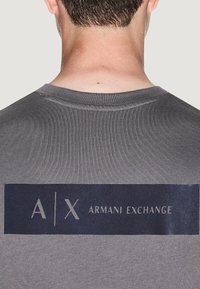 Camiseta gris con un logotipo rectangular en azul en la parte posterior con el texto blanco "A/X ARMANI EXCHANGE". Mangas cortas y diseño de cuello redondo.