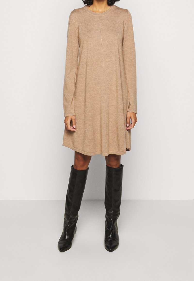 Robe en tricot beige avec un col rond, des manches longues et une silhouette évasée, associée à des bottes en cuir noir à hauteur de genoux.