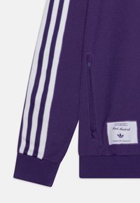 Lila zip-jacka med vita randiga detaljer, ribbade ärmslut och sidoficka. Har en etikett med "Real Madrid" och varumärkesinformation.