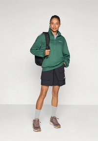 Zelený fleecový svetr s čtvrtinovým zipem, námořnicky modré nákladní šortky s velkými kapsami, šedé ponožky a hnědé turistické boty. Černý batoh přes rameno.