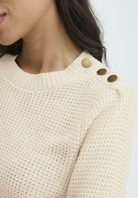 Suéter de punto color crema con cuello acanalado y tres botones decorativos de bronce en el hombro, llevado por una persona con cabello oscuro.