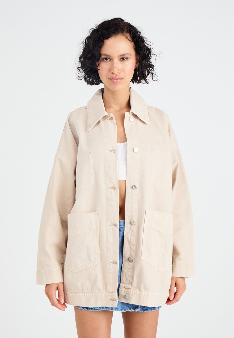 Beige oversized denimjakke med krave, to forlommer og trykknapper. Glat tekstur, afslappet design og løstsiddende pasform.