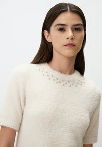 Pull en crème, court à manches courtes, en peluche, avec un col rond décoré d'une rangée de petites embellissements argentés.