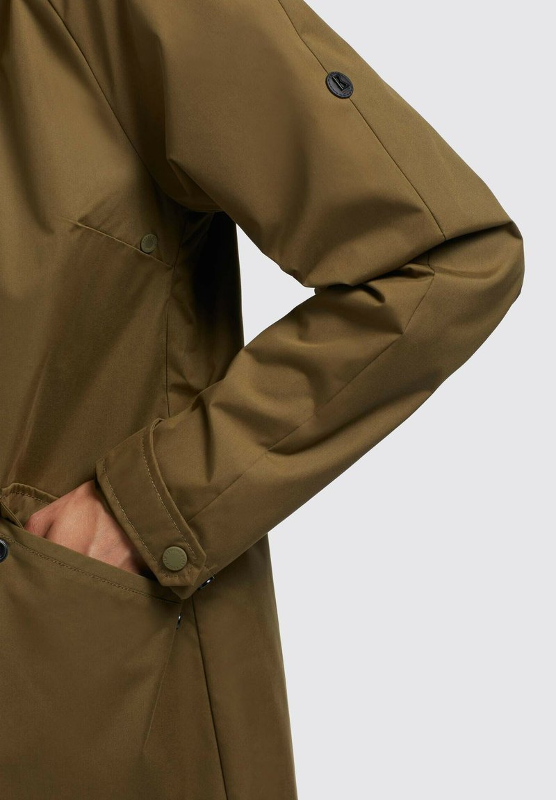 Braune wasserdichte Jacke mit glatter Textur, ausgestattet mit einem Druckknopfverschluss an den Ärmeln und seitlichen Taschen für Funktionalität.