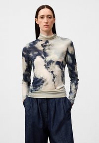 Langsleeve top in beige met blauwe en grijze abstracte patronen, voorzien van een hoge hals en aansluitende manchetten, gecombineerd met donkerdenim broek.