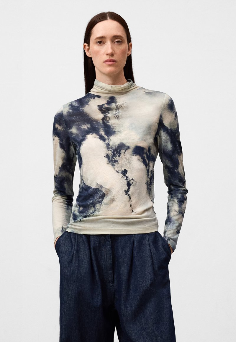 Langsleeve top in beige met blauwe en grijze abstracte patronen, voorzien van een hoge hals en aansluitende manchetten, gecombineerd met donkerdenim broek.