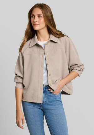 Junge Frau mit langen Haaren, die ein beiges Cordhemd über einem gestreiften Top und blauen Jeans trägt, steht mit einer Hand in der Tasche.