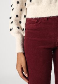 Stickad tröja i kräm med svarta prickar, parat med bordeauxfärgade manchesterbyxor. Tröjan har ballongsärmar och ribbad fåll. Händer synliga.