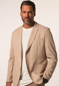 Blazer beige en tissu texturé, avec une poche avant simple et deux boutons. Porté sur un t-shirt blanc, avec une texture lisse.