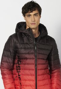 Giubbotto imbottito da uomo con un gradiente dal nero al rosso, texture trapuntata, cappuccio regolabile, zip frontale e dettaglio del logo sul lato.