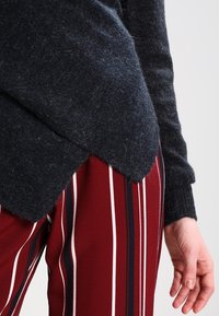 Pull en laine gris foncé avec un ourlet incurvé, associé à un pantalon bordeaux rayé avec des lignes blanches et marine. Gros plan de la main.