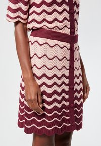 Neulemekko, jossa on vuorottelevia aaltoilevia raitoja burgundissa ja pehmeässä vaaleanpunaisessa, napitus edessä, ribattu vyötärö ja aaltoileva helma.