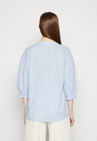 Blusa azul clara em tecido texturizado com mangas bufantes até ao cotovelo e decote redondo. A vista traseira revela detalhes franzidos nos ombros.