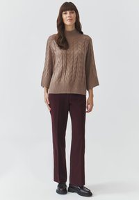 Hellbrauner Kabelstrickpullover mit hohem Kragen, kombiniert mit figurschmeichelnden burgunderfarbenen Hosen; besitzt gerade Beine und schwarze Schuhe.