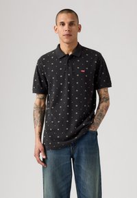 Musta poolpaita, jossa on pieni geometrinen kuvio, kolmen napin listalla ja Levi's-logo. Yhdistetty rentoihin sinisiin farkkuihin.