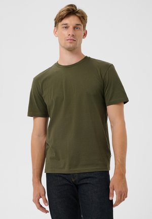 Joven con una camiseta de manga corta verde oliva y vaqueros oscuros, de pie sobre un fondo claro y uniforme.