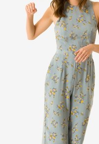 Lyseblå floral jumpsuit med en tætsiddende overdel og bukser med vide ben, med gule og grå blomsterprint på et blødt stof.