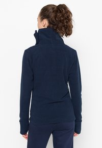 Giacca in pile blu navy con colletto alto, maniche lunghe e una texture morbida. Presenta un design aderente e senza hardware o logo visibili.