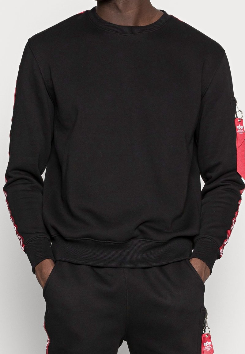 Homme portant un sweat-shirt noir et un pantalon avec des bandes rouges et des tirettes de fermeture éclair rouges sur les manches et les poches, les mains dans les poches.