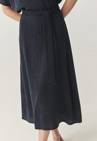 Donna che indossa una gonna midi blu navy con una cintura annodata in vita, in piedi con le mani dietro la schiena su uno sfondo chiaro.