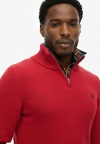 Suéter de punto rojo con un cuello alto y cremallera, con un patrón de cuadros en el cuello y un logotipo en el pecho.