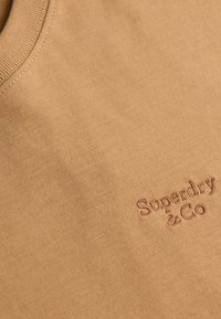 Gros plan sur un tissu beige avec « Superdry & Co » brodé avec un fil assorti près d’un col côtelé.