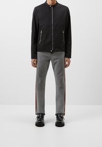 Zwarte ritssluiting jack met twee voorzakken en staande kraag, gecombineerd met grijze jeans met een zijbrown streep en zwart schoeisel.