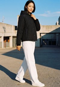 Jonge vrouw in een zwarte doorgestikte jas en witte broek die op een zonnige dag buiten wandelt met moderne betonnen gebouwen op de achtergrond.