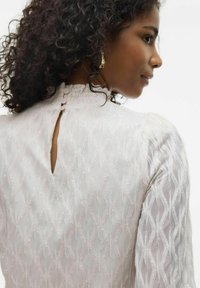 Witte textuurblouse met een golvend patroon, gerimpelde kraag en een sleutelgat achteraan met knoopsluitingen. Krullend haar en gouden oorbellen zichtbaar.