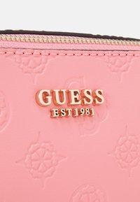 Lyserød lædervare med præget blomsterdesign, gylden lynlås og gyldent "GUESS EST 1981" logo på fronten.